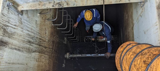 Two workers wearing safety gear and hardhats are repairing pipes in a deep trench