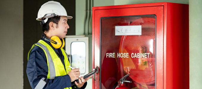 A safety inspector in a hard hat and high-visibility vest stands next to a red wall-mounted fire hose cabinet, holding a tablet and pen to conduct a routine check.