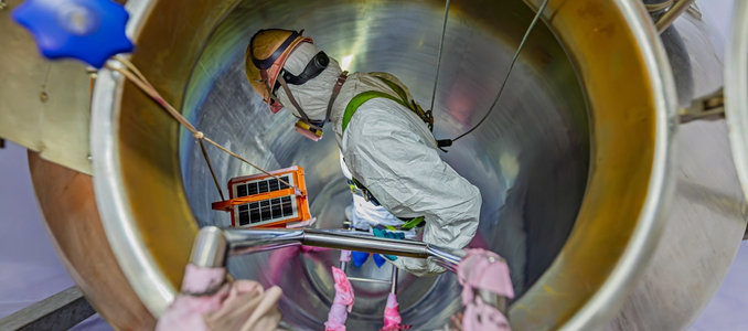 A worker wearing full protective suit, helmet, respirator, and safety harness inspects the interior of a large, cylindrical metal tank.