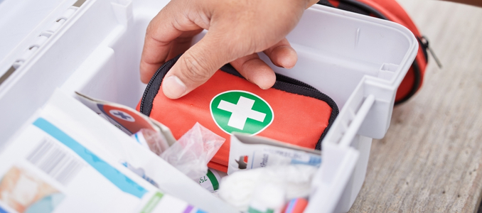 A hand reaching into an open white plastic first aid box to grab a small red pouch with a green first aid cross emblem.