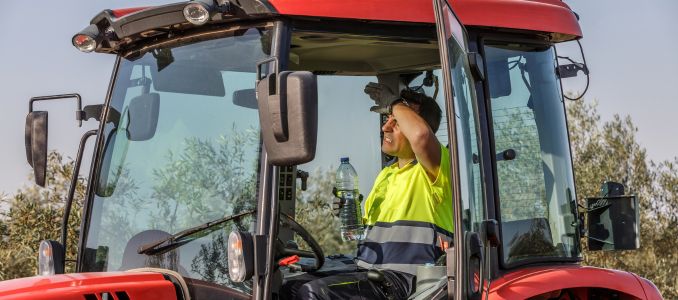Agricultural worker wearing a high-visibility shirt wipes his forehead while holding a water bottle inside a tractor cab.