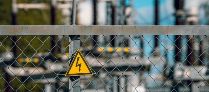 A yellow high-voltage warning sign attached to a chain-link fence at an electrical substation.
