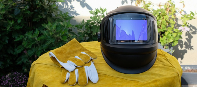 An auto-darkening welding helmet with a digital screen alongside heavy-duty leather welding gloves resting on a yellow cloth surface.