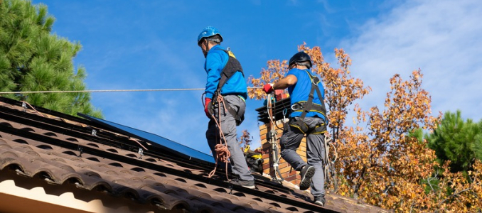 Two workers wearing safety helmets and harnesses attached to fall protection lifelines installing solar panels on a tiled roof.
