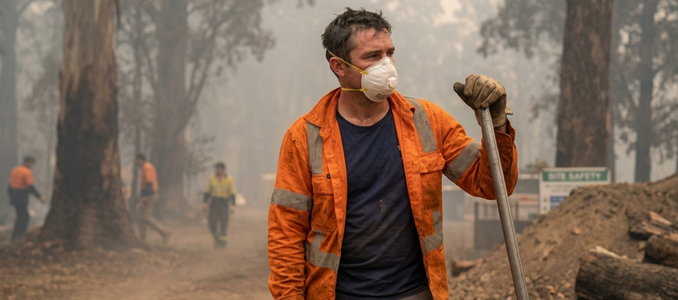 A worker wearing a white N95 mask and an orange high-vis jacket stands in a smoky forest, leaning on a tool handle with other workers in the background.