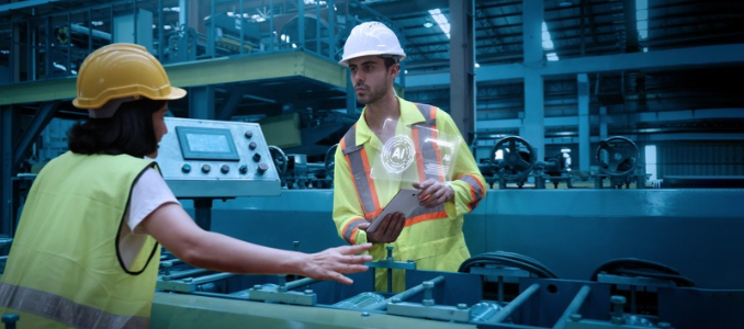 Two factory workers wearing hard hats and safety vests collaborating at an industrial machine, with one holding a tablet displaying artificial intelligence graphics.