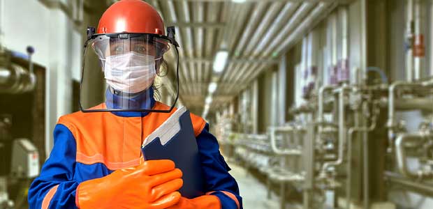 person wearing hard hat, face shield, face mask, gloves and protective shirt while holding a clipboard in a factory