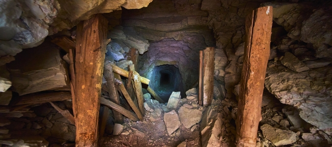 Interior view of a dimly lit underground mine shaft framed by weathered wooden support beams leading into a dark tunnel.