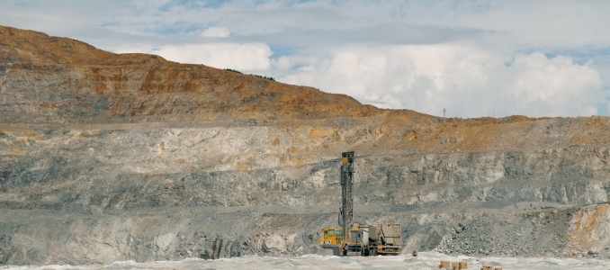 A large, yellow blast-hole drilling rig stands on the floor of a massive open-pit mine, set against a backdrop of tiered rock faces and a cloudy sky.