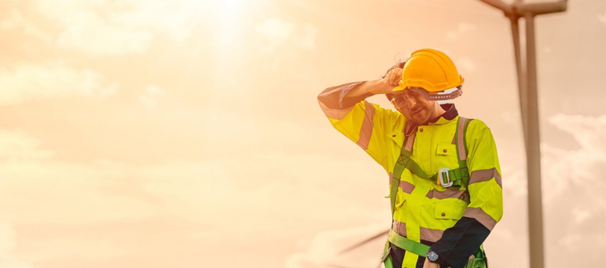 An outdoor worker wearing a hard hat and high-visibility clothing wipes sweat from his forehead under a bright, hot sun.