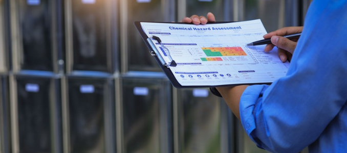A person in a blue shirt holding a clipboard titled "Chemical Hazard Assessment" and writing on it with a pen.