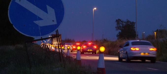 Traffic driving past a line of illuminated traffic cones and road signs along a highway construction zone at dusk.