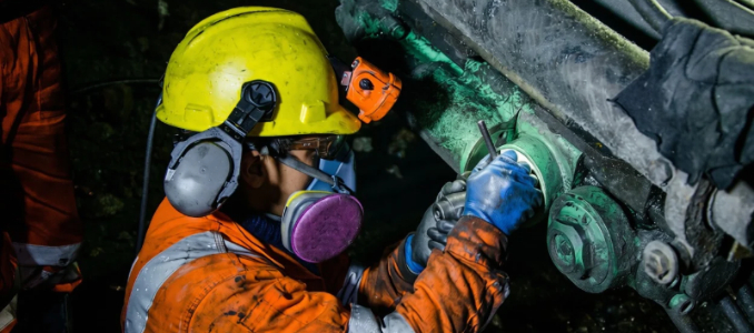 A miner wearing a yellow hard hat with a headlamp, earmuffs, and a respirator mask carrying out maintenance work on heavy equipment underground.