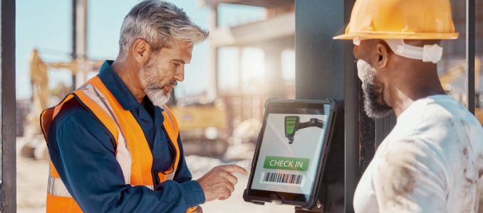 Worker in a safety vest uses a digital check-in kiosk while another worker wearing a hard hat looks on.