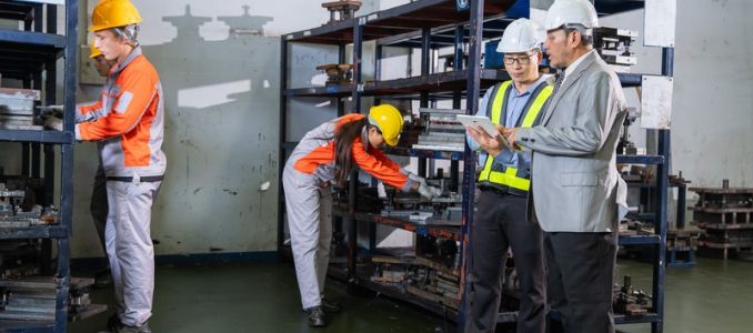 Two workers in hard hats review a tablet while two others handle materials on metal shelving in an industrial facility.